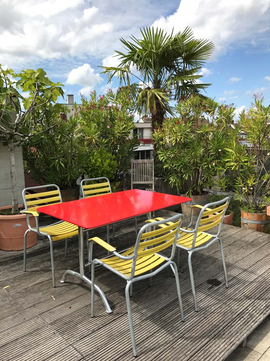 Roter Bättig Gartentisch mit Bättig Gartenstühlen in Zitronengelb auf einer Dachterrasse mit Palmen im Hintergrund