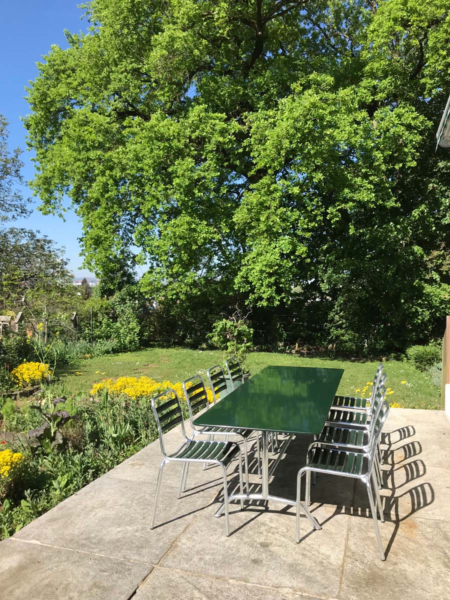 Langer Bättig Gartentisch mit acht Bättig Gartenstühlen in Tannengrün, in der Sonne in einem Garten