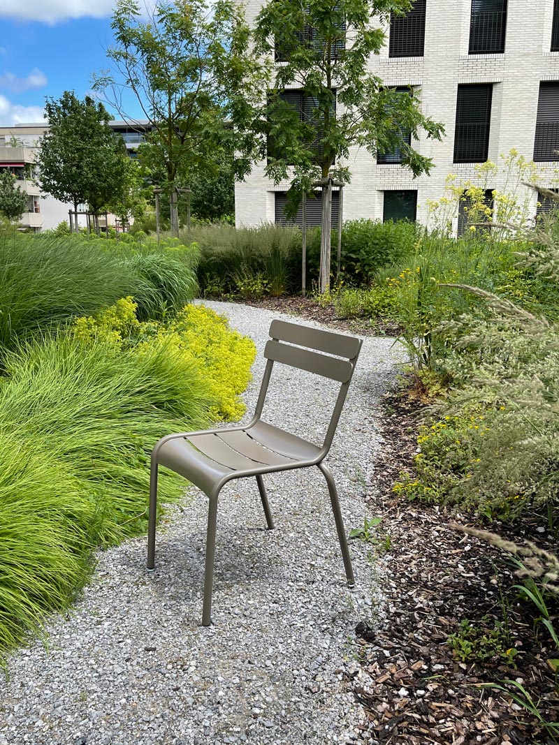 Luxembourg Gartenstuhl in der Sonderfarbe «Schlamm» auf einem Kiesweg in einem grünen Garten.