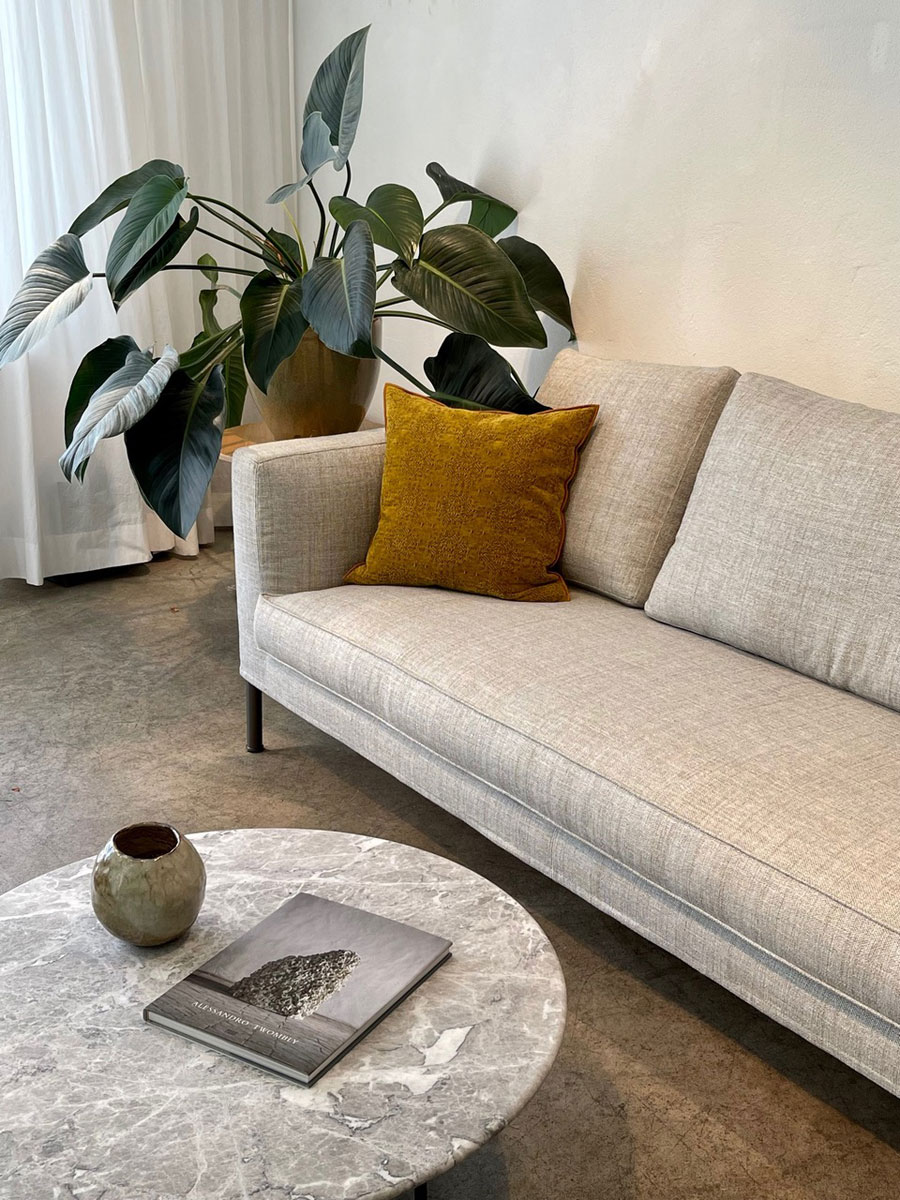 Tiziano Sofa in hellgrau mit ockerfarbenem Kissen. Salontisch mit Vase und Buch im Vordergrund.