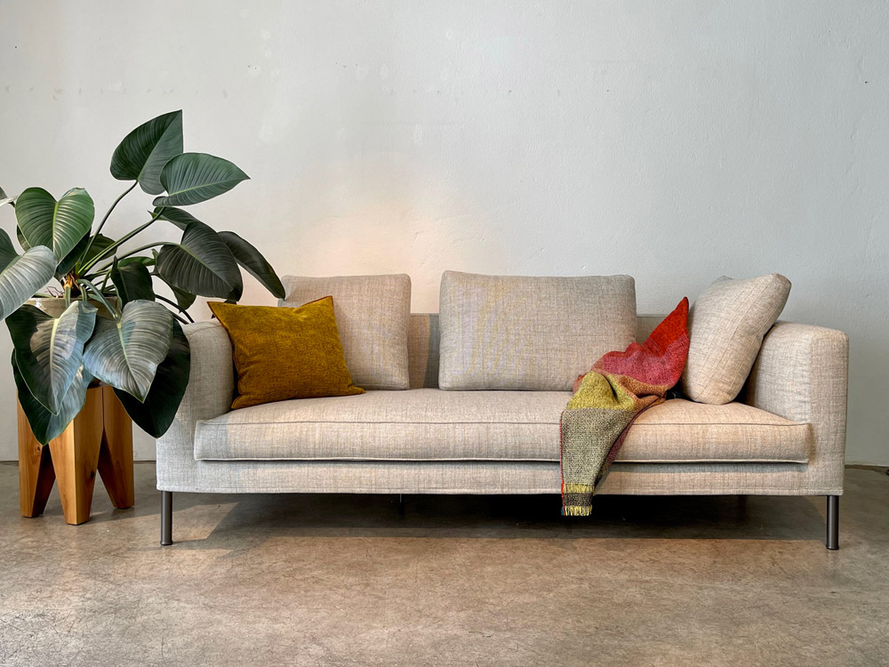 Frontale Ansicht des Tiziano Sofas in hellgrau. In der linken Ecke liegt ein ockerfarbenes Kissen, rechts eine drapierte rotgelbe Decke.