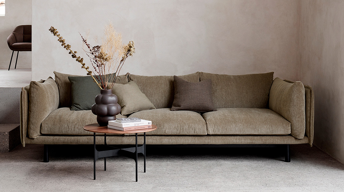 Frontale Aufnahme des Kite Sofas von Wendelbo, mit drei Sitzkissen. Vor dem Sofa ein kleiner, runder Tisch mit Trockenblumen und Büchern.