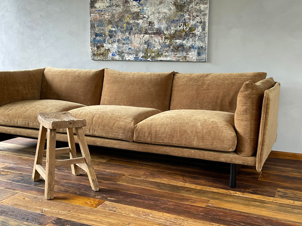 Frontale Aufnahme des Kite Sofas von Wendelbo, mit drei Sitzkissen und gelbem Stoffbezug. Im Hintergrund hängt eine abstrakte Malerei an der Wand, vor dem Sofa steht ein Hocker aus altem Holz.